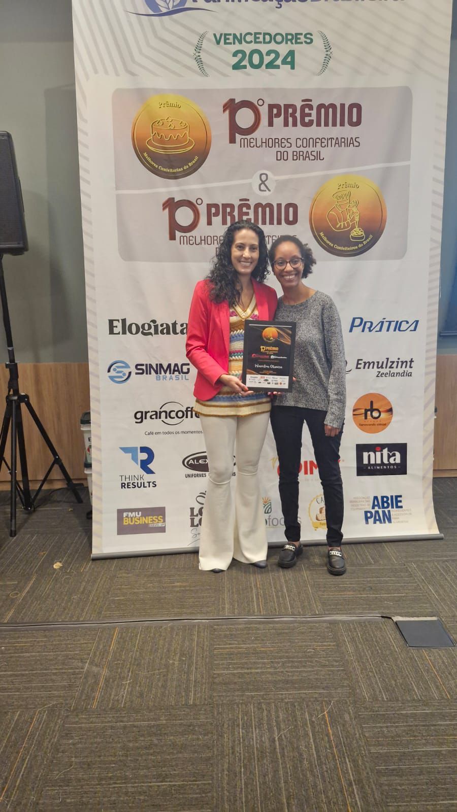 Duas mulheres estão de pé, uma ao lado da outra, em frente a uma parede, segurando um troféu.