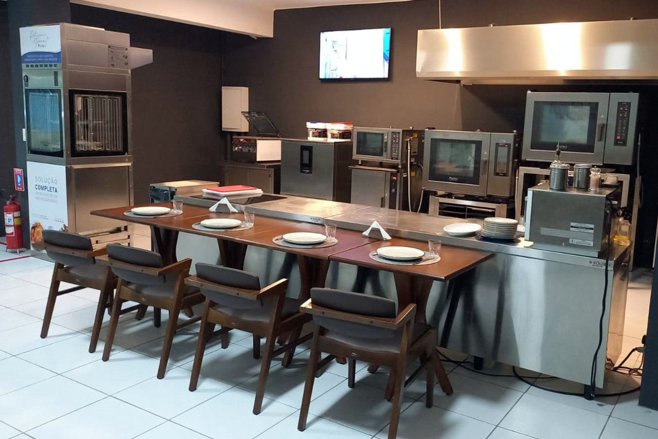 Uma cozinha com uma mesa longa, cadeiras e muitos eletrodomésticos.