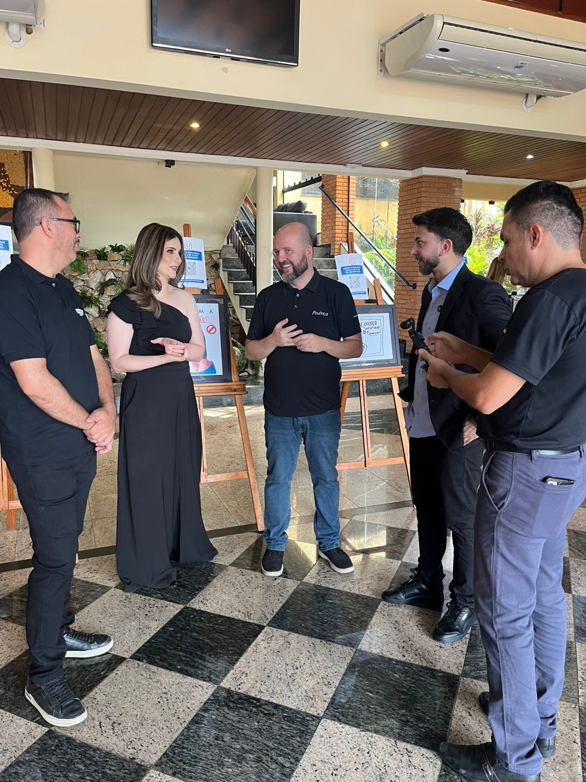 Grupo de pessoas, evento formal, em pé, em ambiente interno. Mulher de vestido preto, homens em trajes casuais, alguns sorrindo.