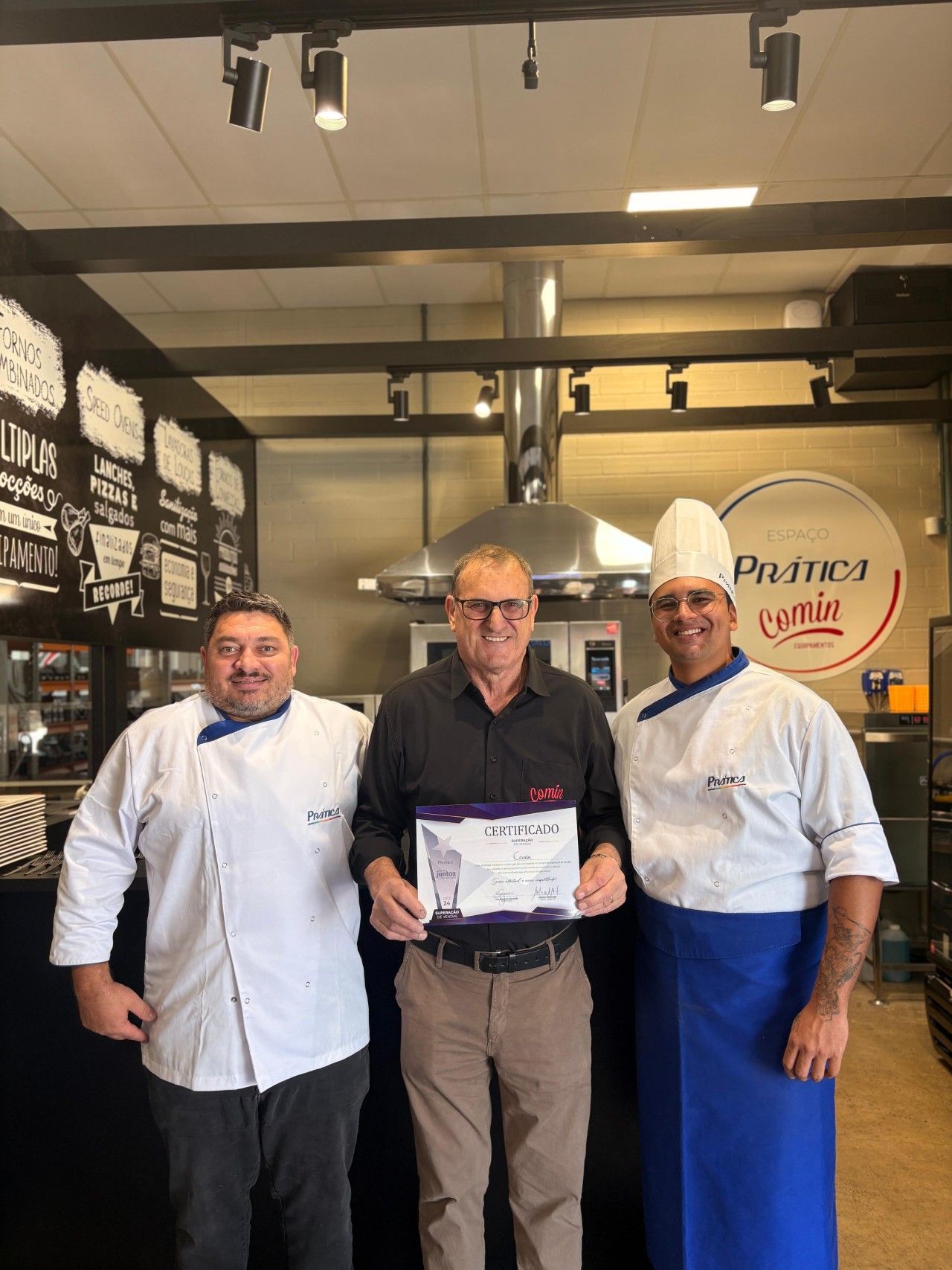 Três homens estão de pé, um ao lado do outro, em uma cozinha, segurando um certificado.