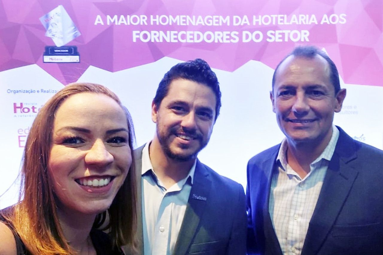 Três pessoas posam para uma foto em frente a uma placa que diz a maior homenagem da hotelaria