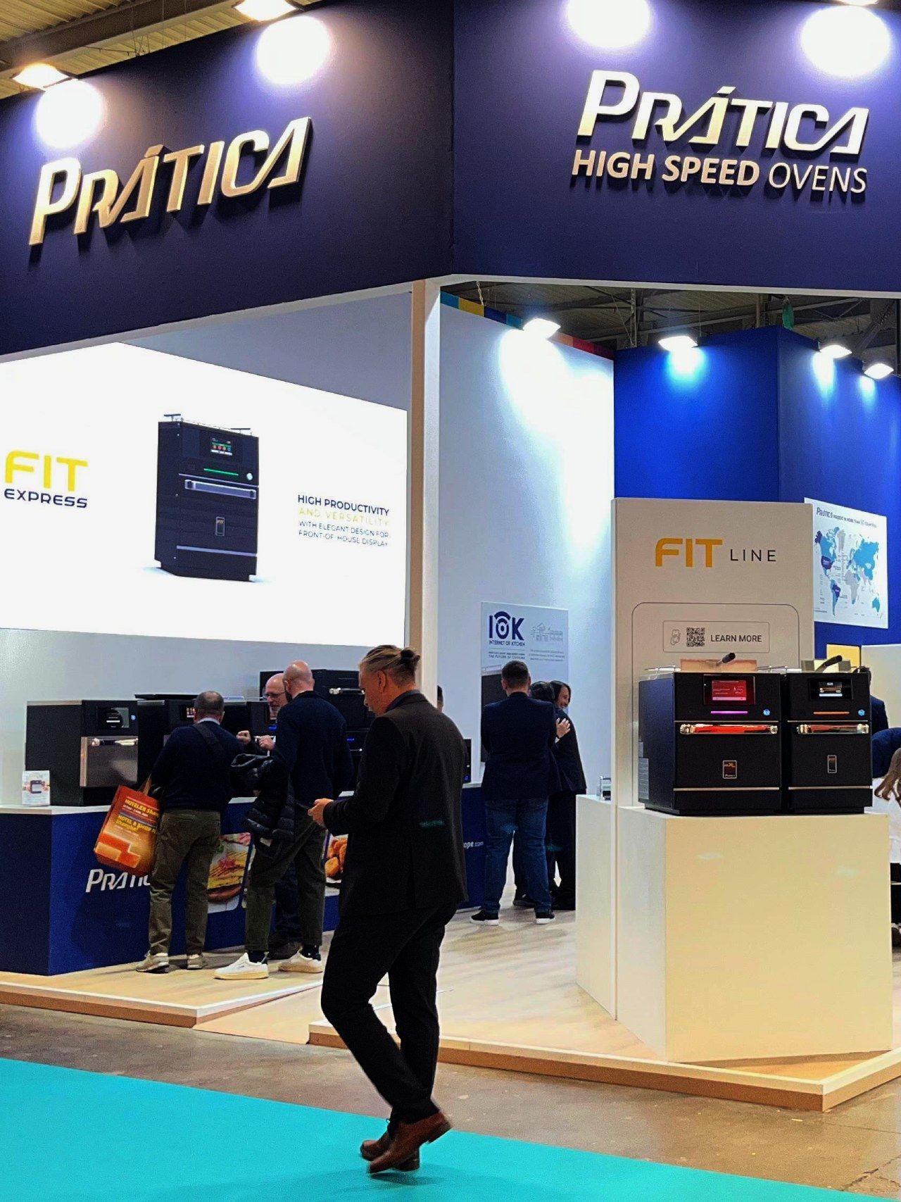 Estande da Prática sobre fornos de alta velocidade com pessoas; marca azul e branca, fornos em exposição, homem passa.