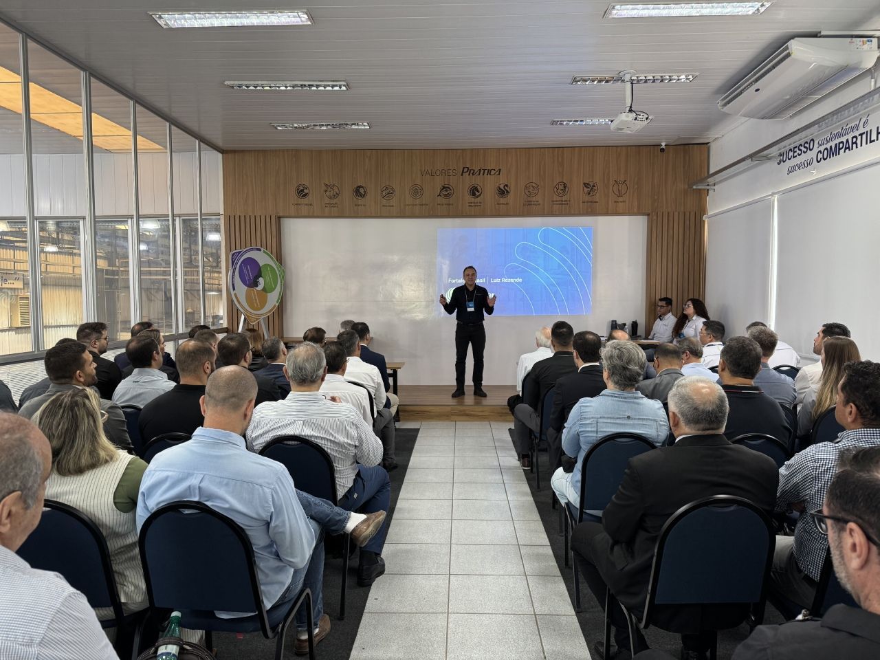 Um homem faz uma apresentação em uma conferência. Os participantes sentam-se em fileiras. Uma tela exibe um fundo azul.