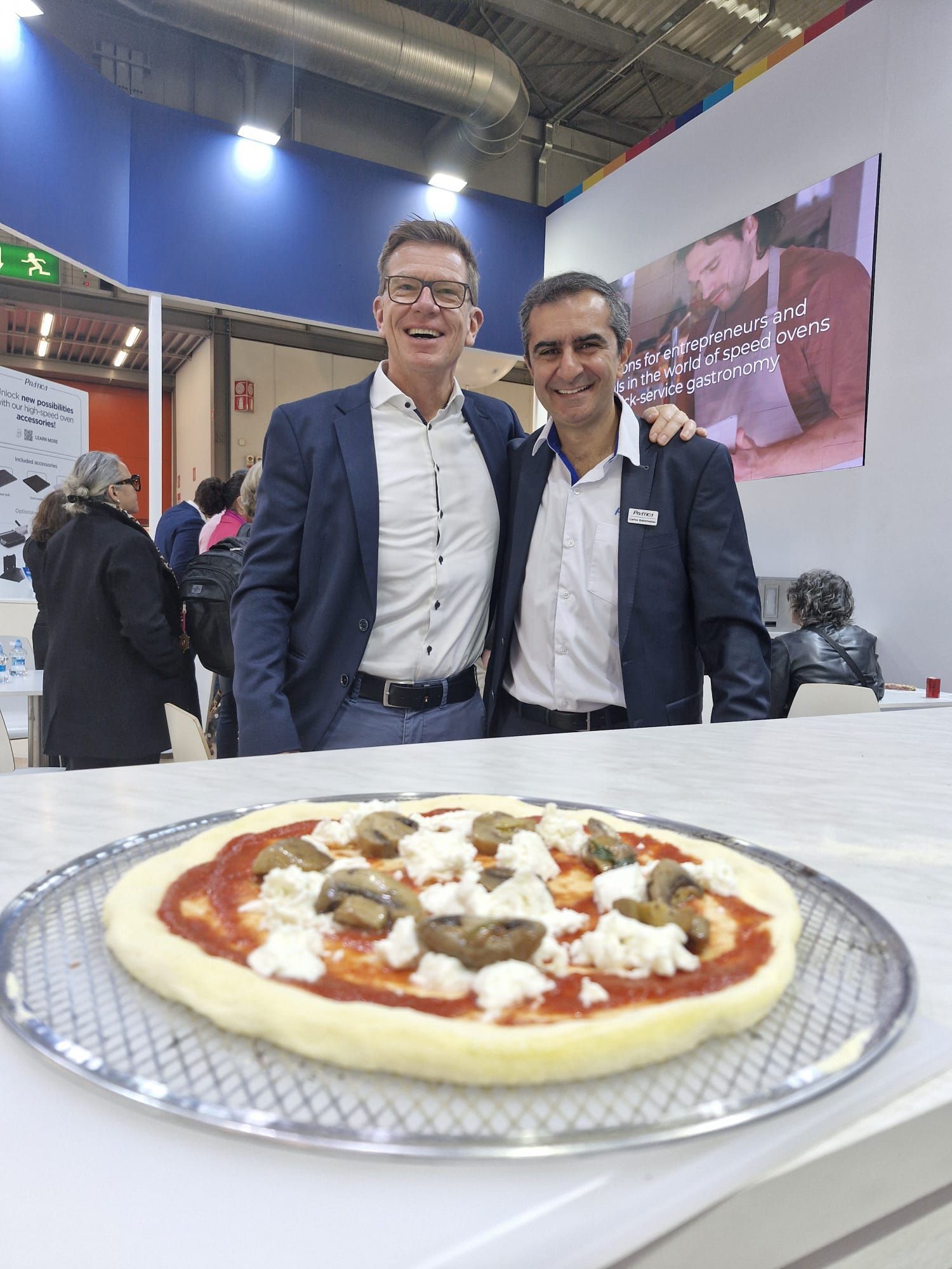 Dois homens posam ao lado de uma pizza com cogumelos e mussarela. Bancada branca, ambiente interno.