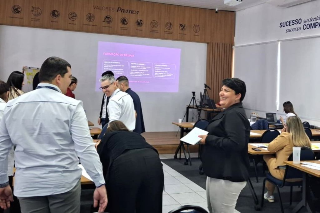 Pessoas em uma sessão de treinamento. Algumas em pé, conversando e sorrindo. Outras sentadas em mesas. Sala de cor clara com tela.