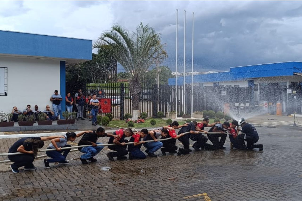 Bombeiros praticam com mangueira, ajoelhando-se e lançando água perto de um prédio azul, sob o olhar atento de espectadores.