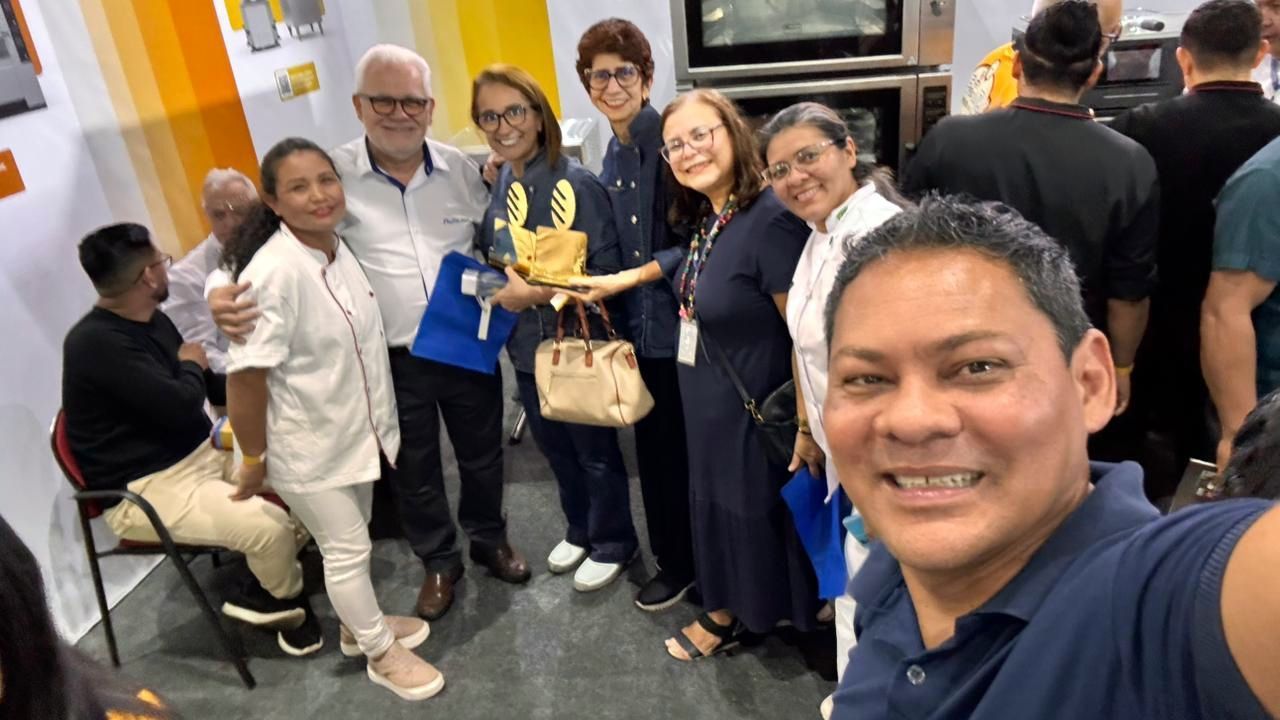 Selfie em grupo em um estande de feira comercial, pessoas sorridentes posando com um homem tirando a foto.
