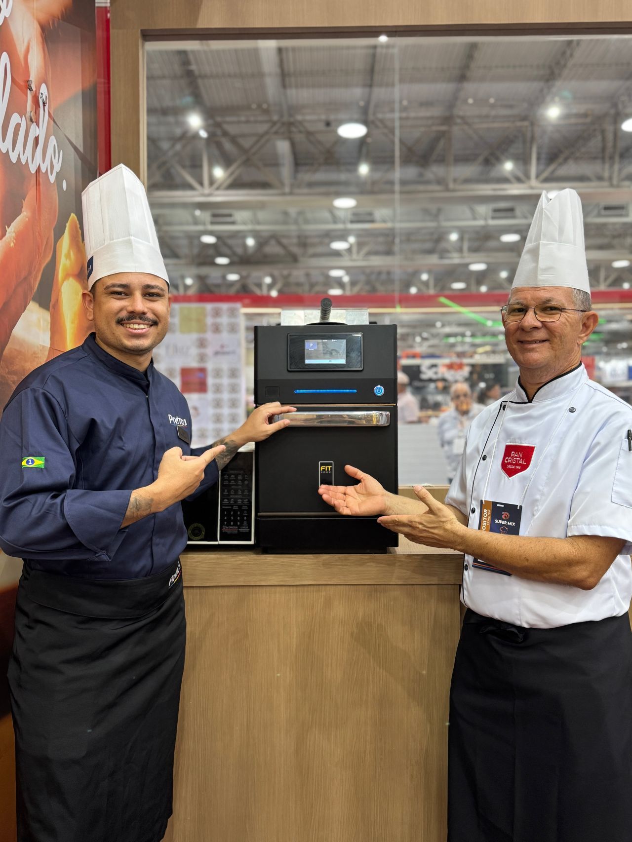 Dois chefs de chapéu branco apontam para um forno preto com uma tela em uma feira gastronômica.
