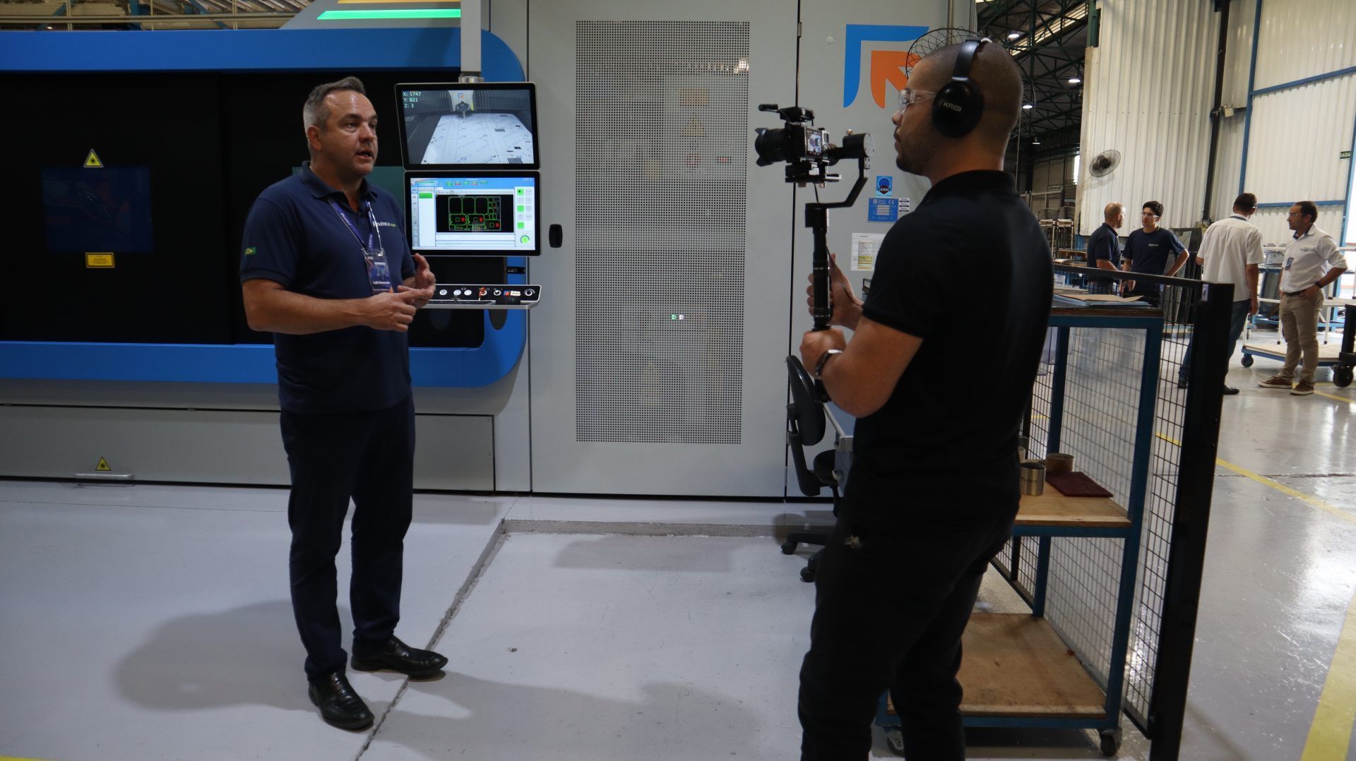 Um homem está em frente a uma câmera em uma fábrica conversando com outro homem.