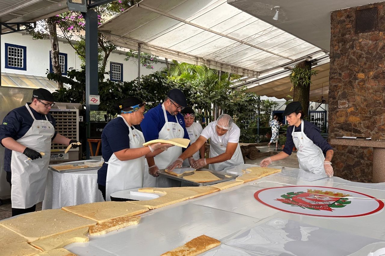 Grupo de pessoas montando camadas de bolo em uma grande mesa ao ar livre. Os chefs usam aventais e estão concentrados no preparo dos alimentos.