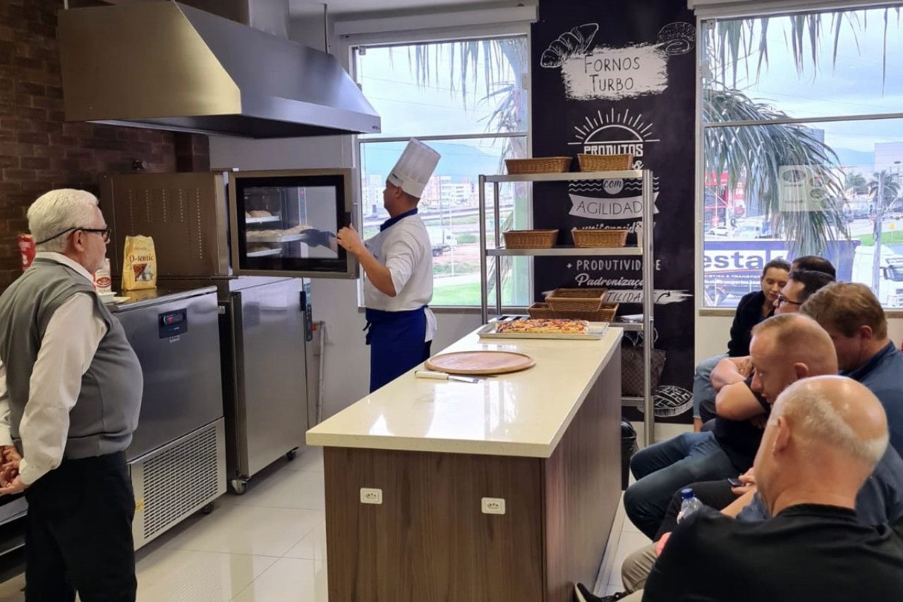 Um homem com chapéu de chef está parado na frente de um grupo de pessoas em uma cozinha.