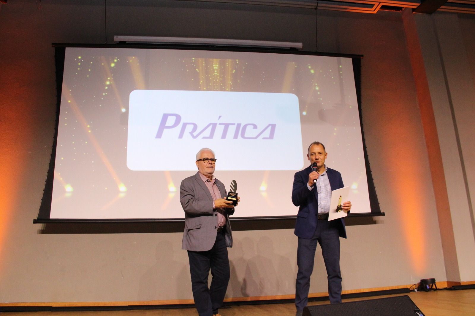 Dois homens estão em pé diante de uma tela grande com a palavra pratica escrita nela.