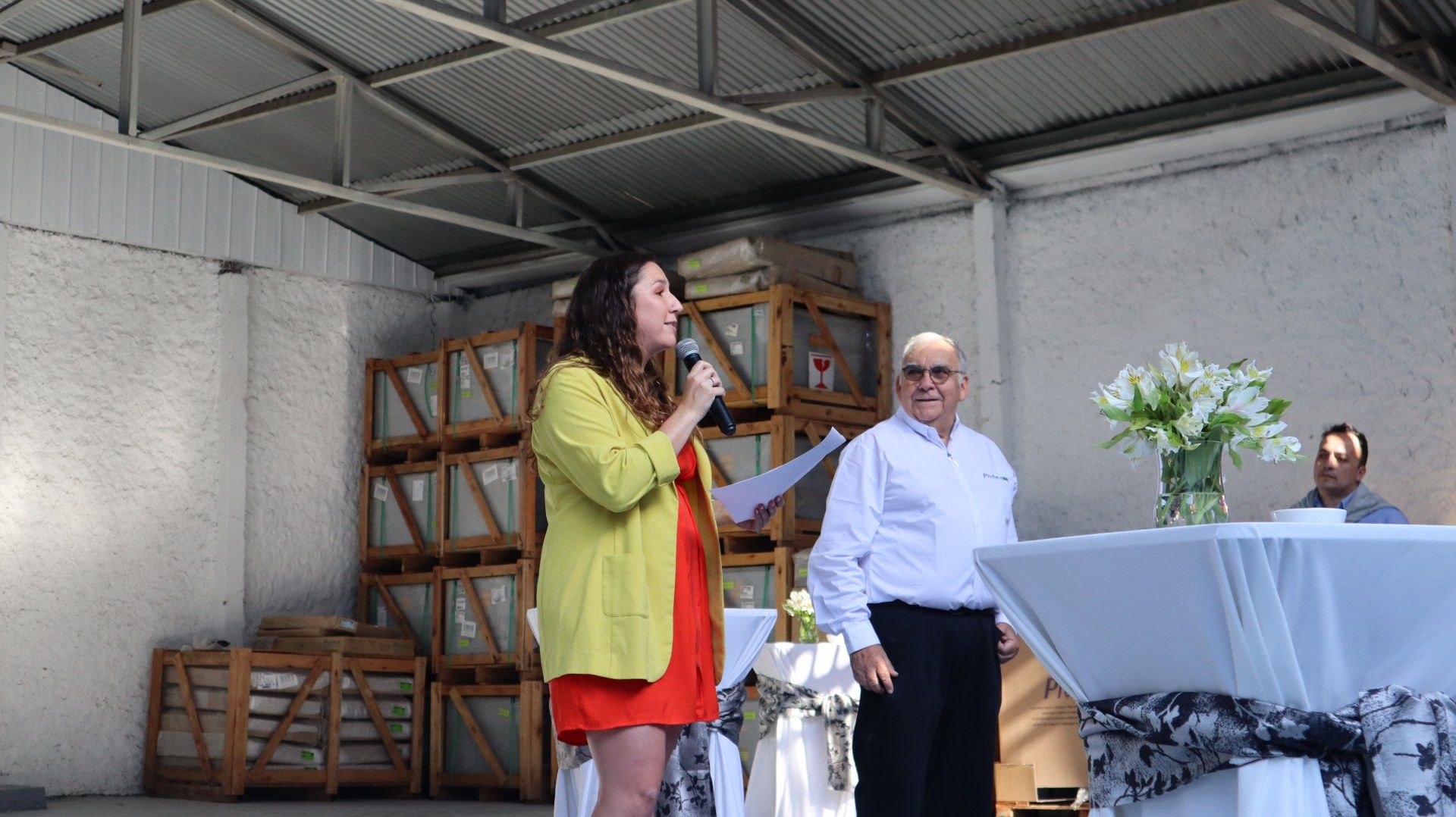 Uma mulher está falando em um microfone enquanto está ao lado de um homem em um armazém.