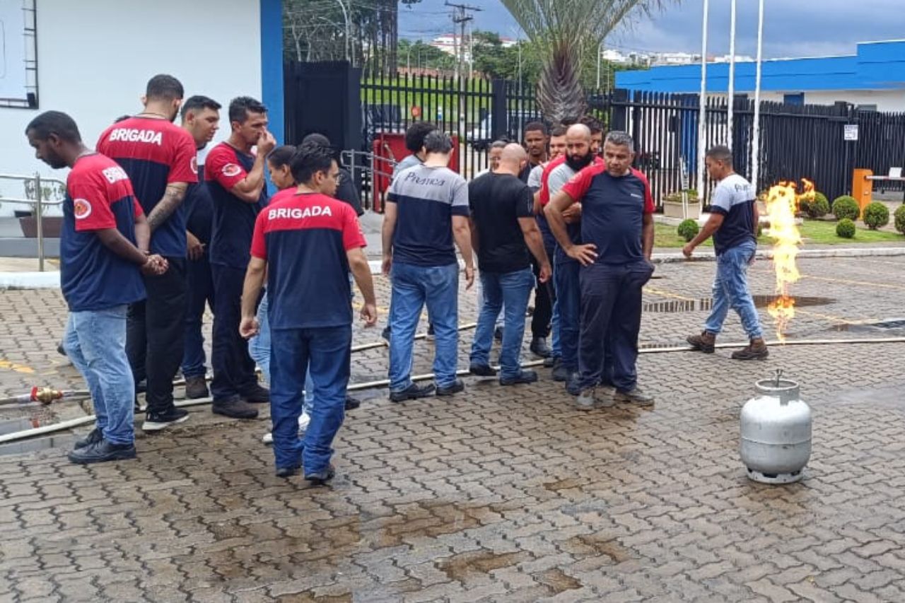 Grupo de pessoas observando um incêndio em um tanque de propano em um ambiente externo. Alguns vestem camisetas de uniforme vermelhas e azuis.