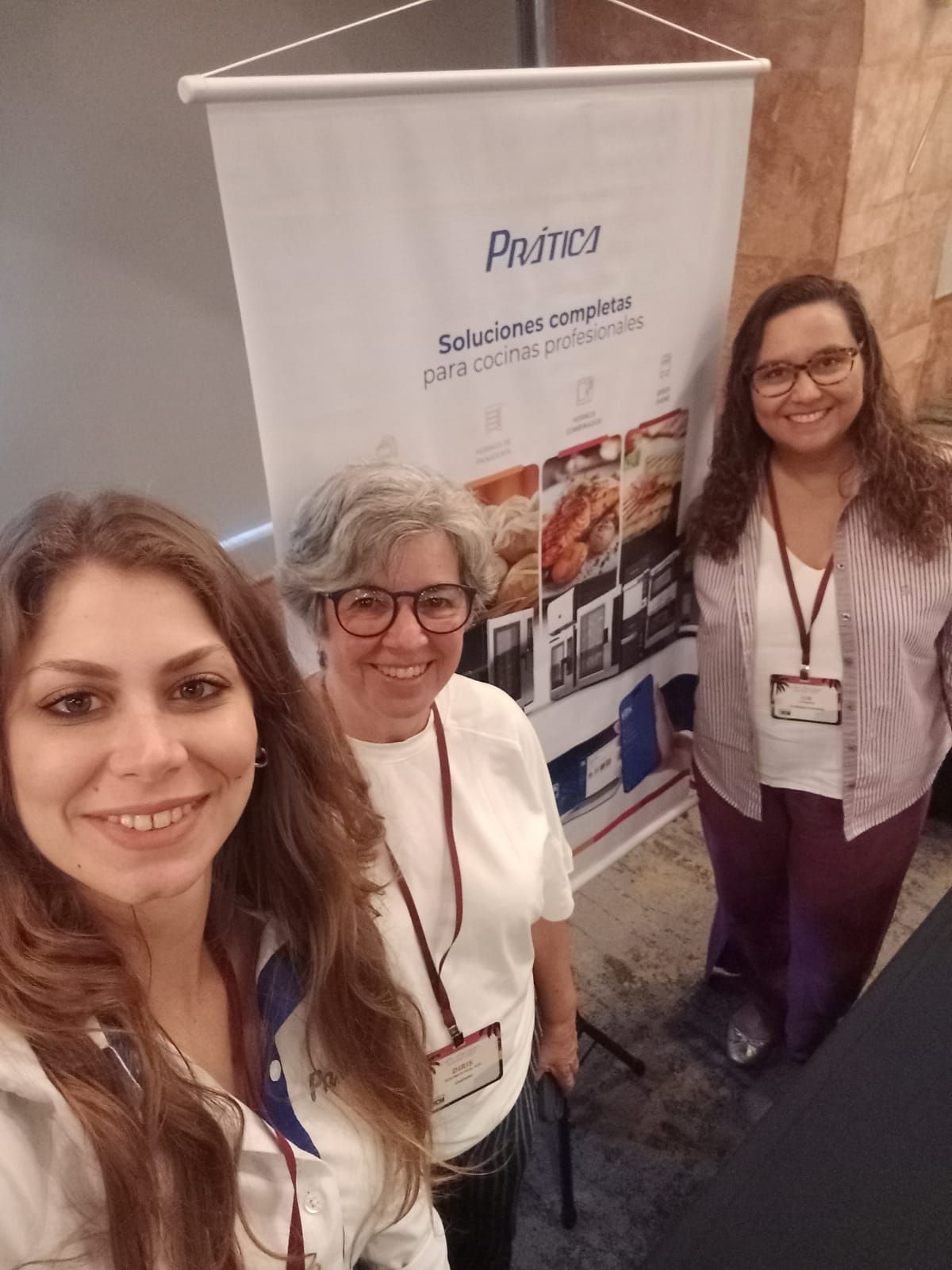 Três mulheres estão em frente a uma faixa. Duas sorriem, uma tira uma selfie. A faixa é da