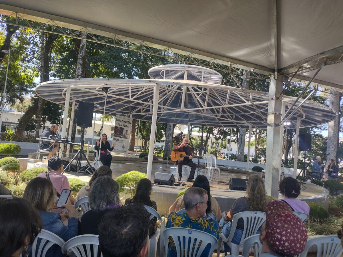 Um grupo de pessoas está sentado sob uma tenda observando um homem tocar violão.