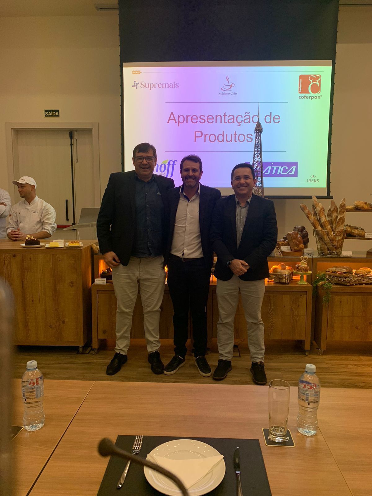 Três homens posam para uma foto em frente a uma tela de projetor.