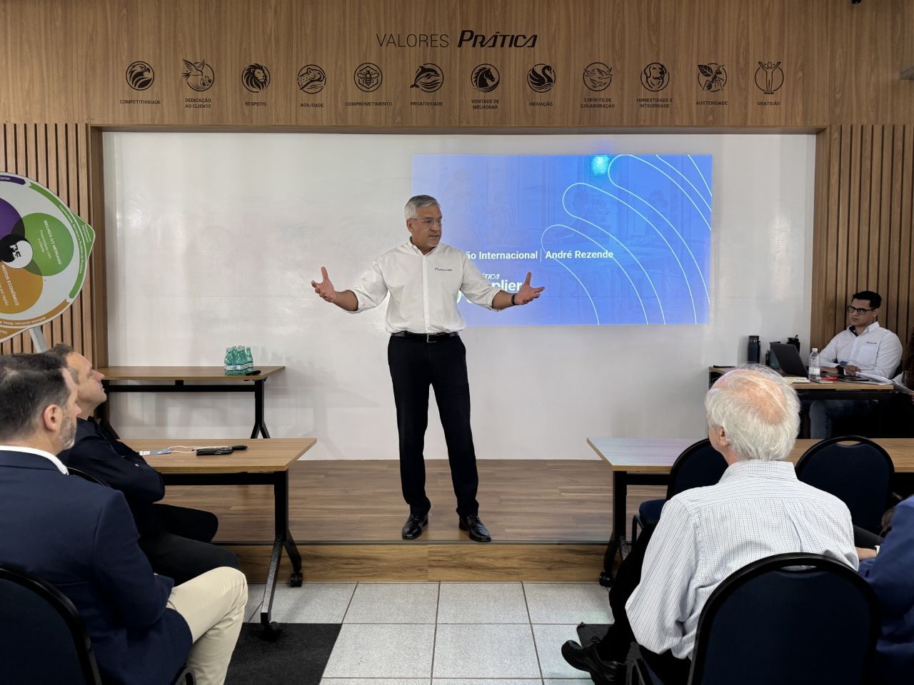 Homem apresentando para uma plateia em ambiente fechado, em pé perto de uma tela. Paredes de madeira, mesas e pessoas sentadas.