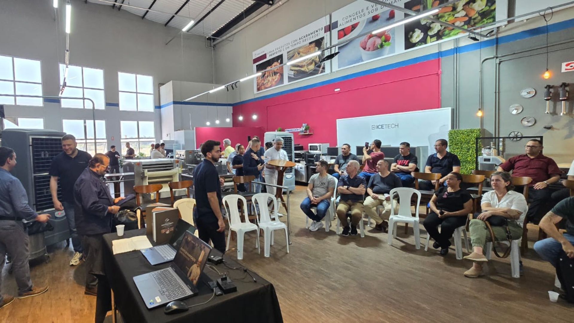 Pessoas em um ambiente de oficina, ouvindo uma apresentação. Sala ampla com cadeiras, mesas e utensílios de cozinha.