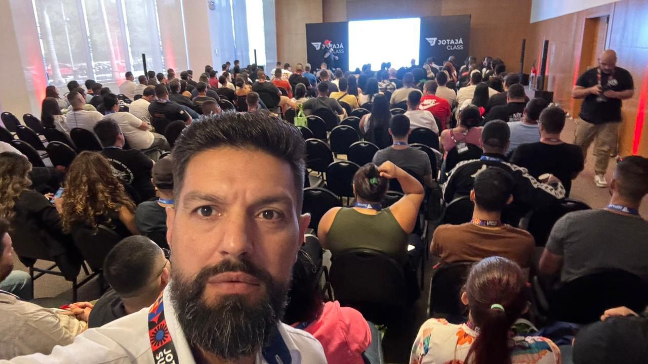 Um homem com barba está tirando uma selfie na frente de uma multidão em uma sala de conferências.