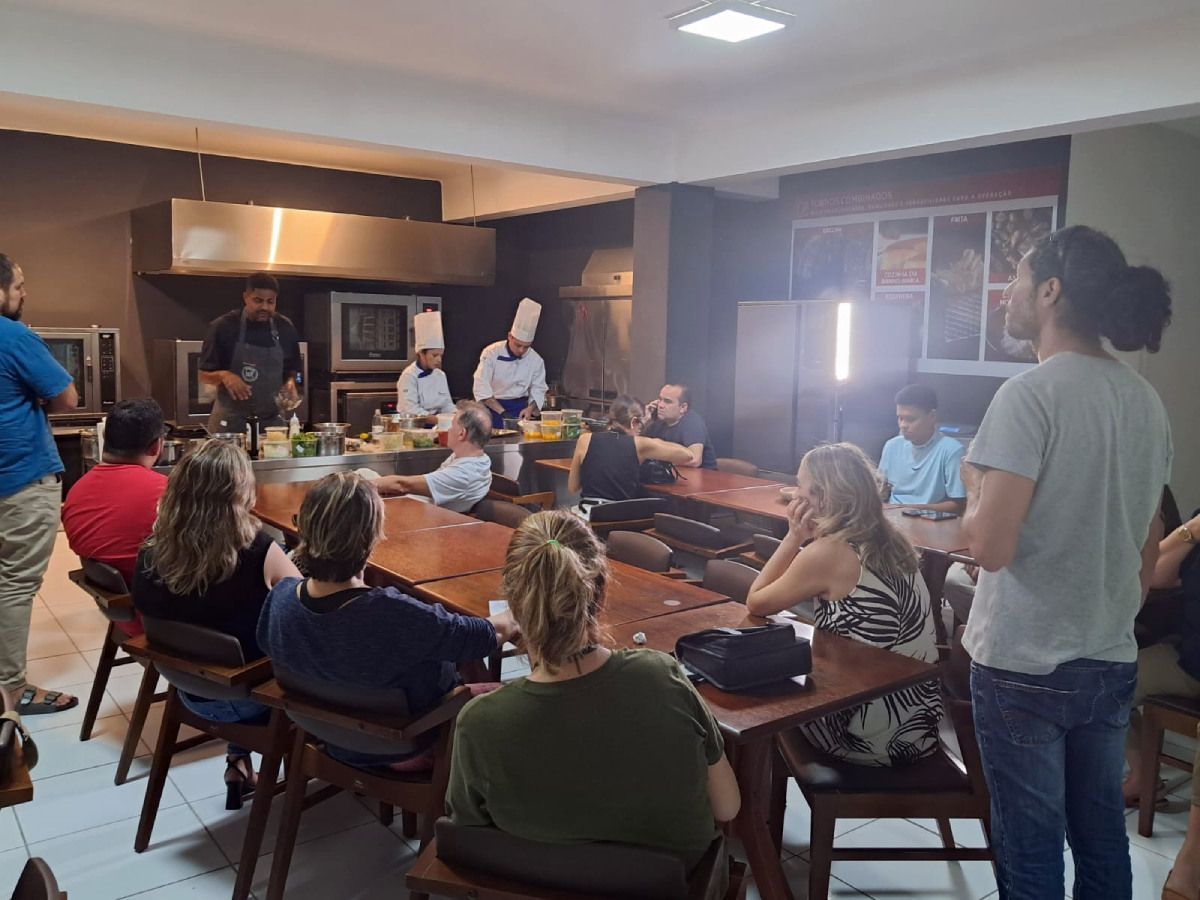 Um grupo de pessoas está sentado em mesas em uma sala observando um chef cozinhar.