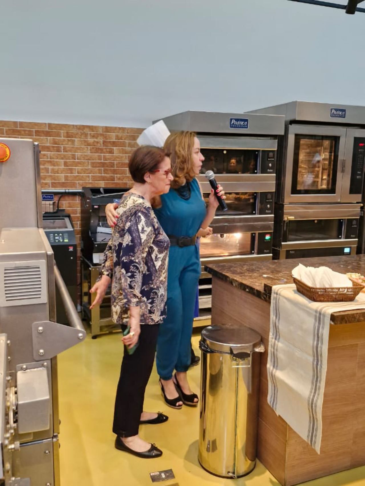 Duas mulheres estão de pé, uma ao lado da outra, em uma cozinha, com um microfone.