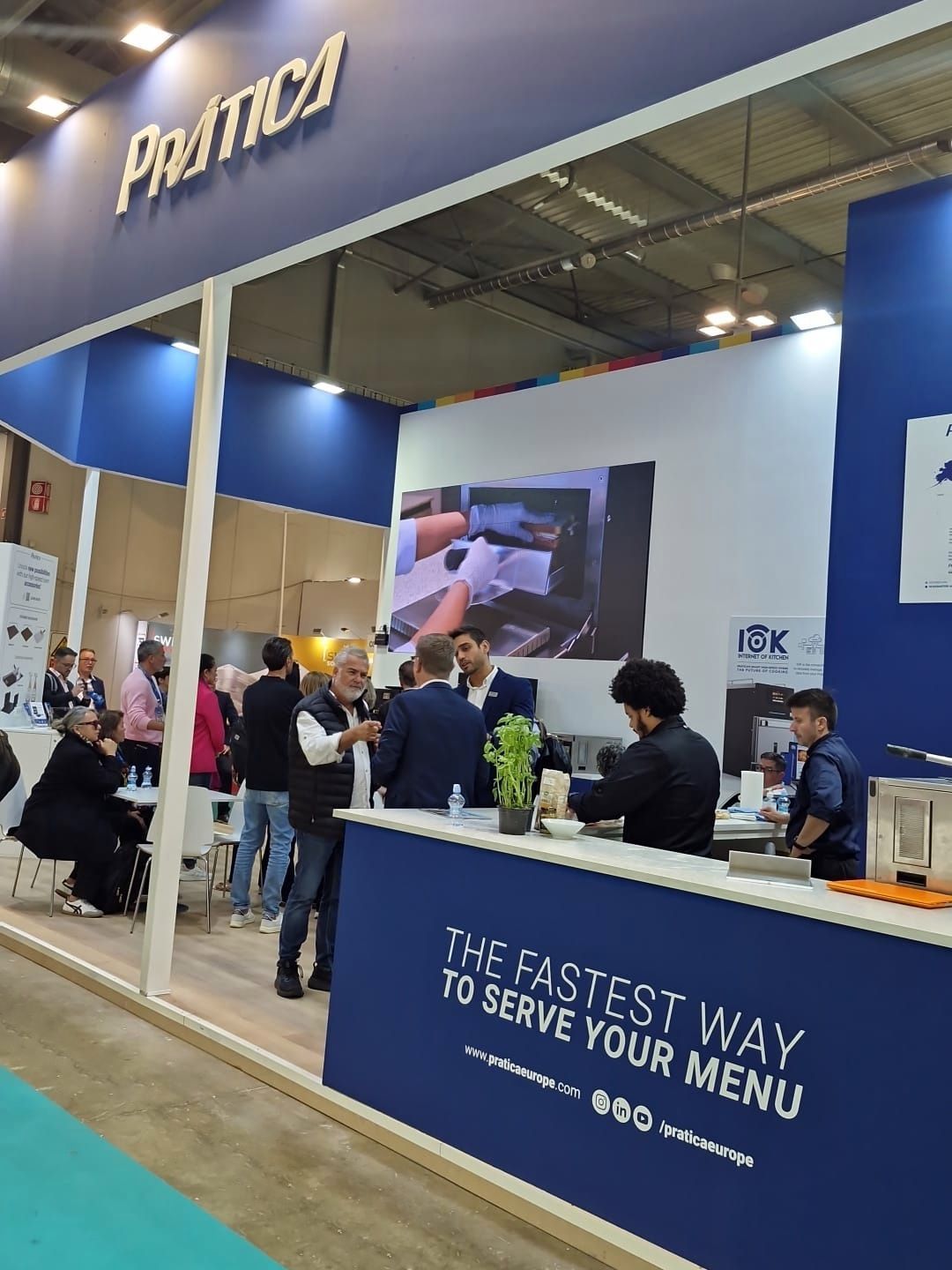 Estande da Prática em uma feira comercial: pessoas em networking, cores branco e azul, texto 