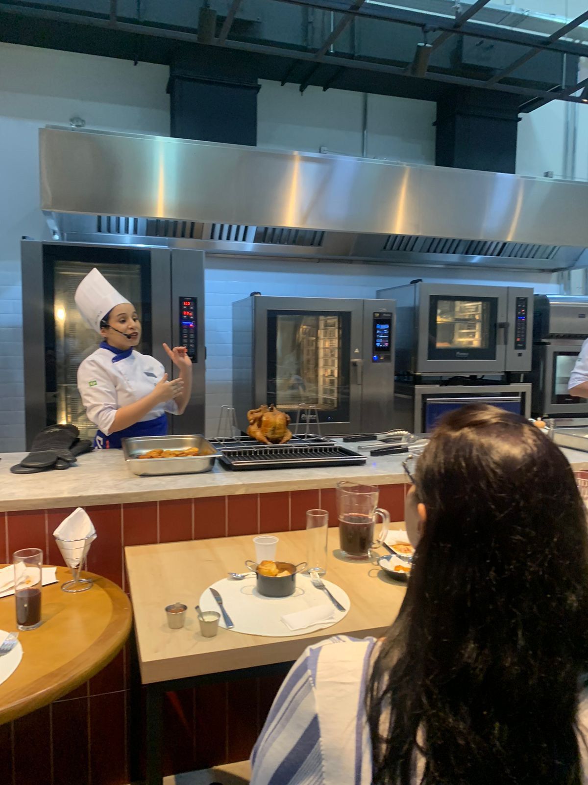 Uma mulher está sentada à mesa em frente a um chef em uma cozinha.