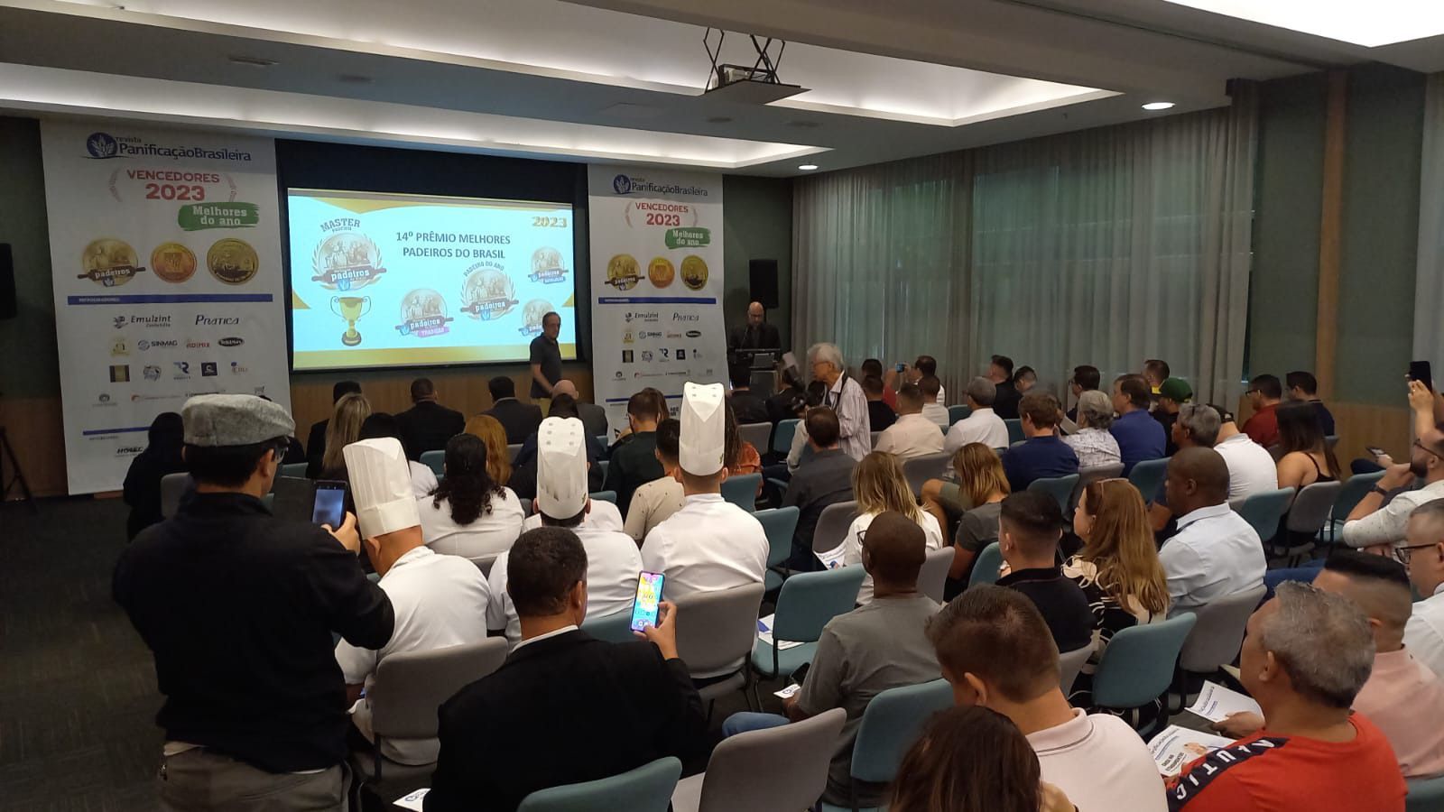 Um grande grupo de pessoas está sentado em uma sala de conferências assistindo a uma apresentação.