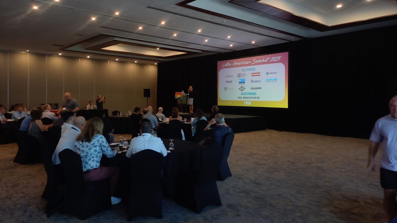 Conferência com pessoas sentadas em mesas, apresentador no palco com um slide mostrando logotipos.