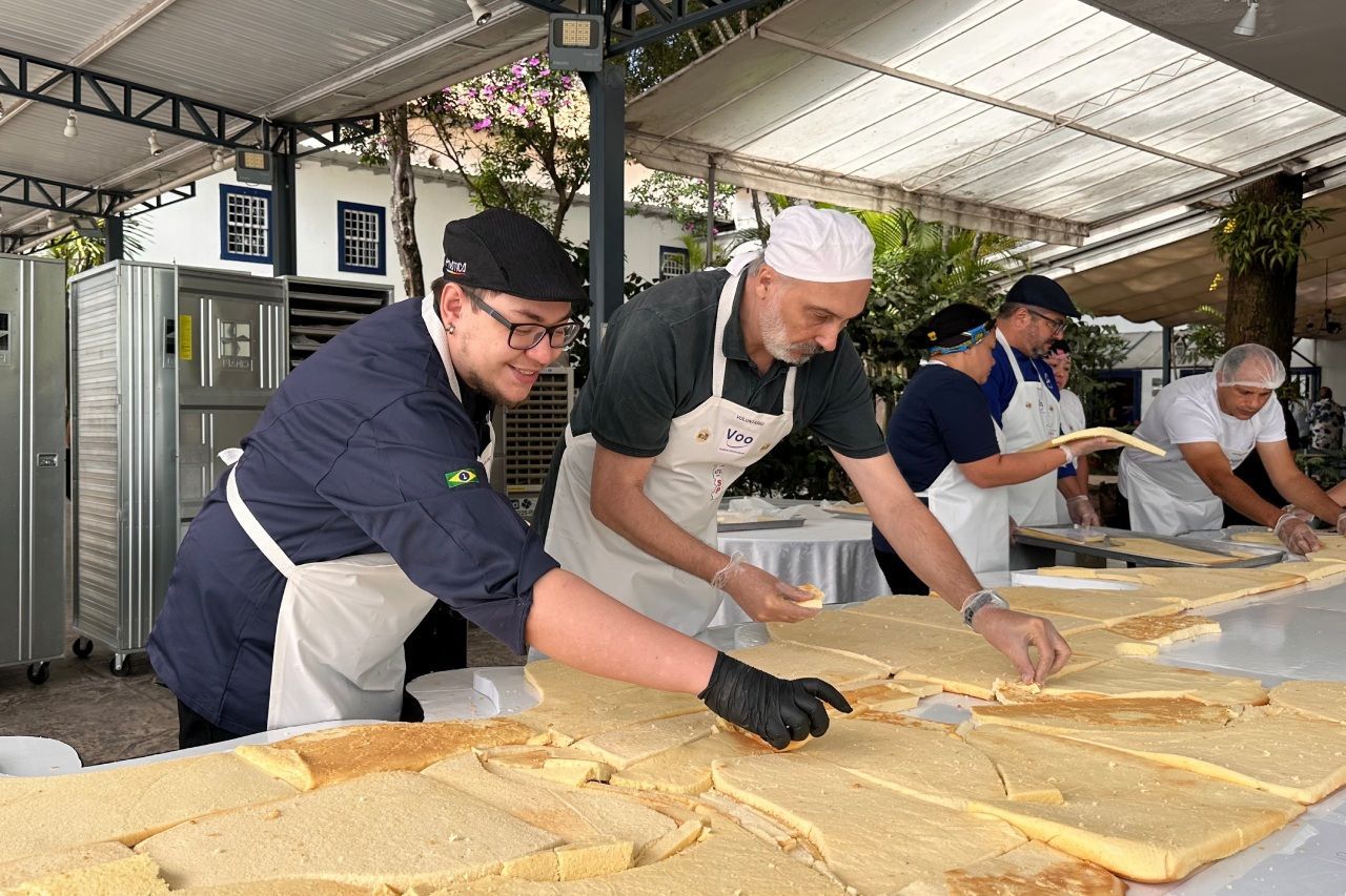 Pessoas preparando uma grande fatia de comida ao ar livre. Os trabalhadores usam aventais e chapéus, fatiando-a em uma mesa comprida.