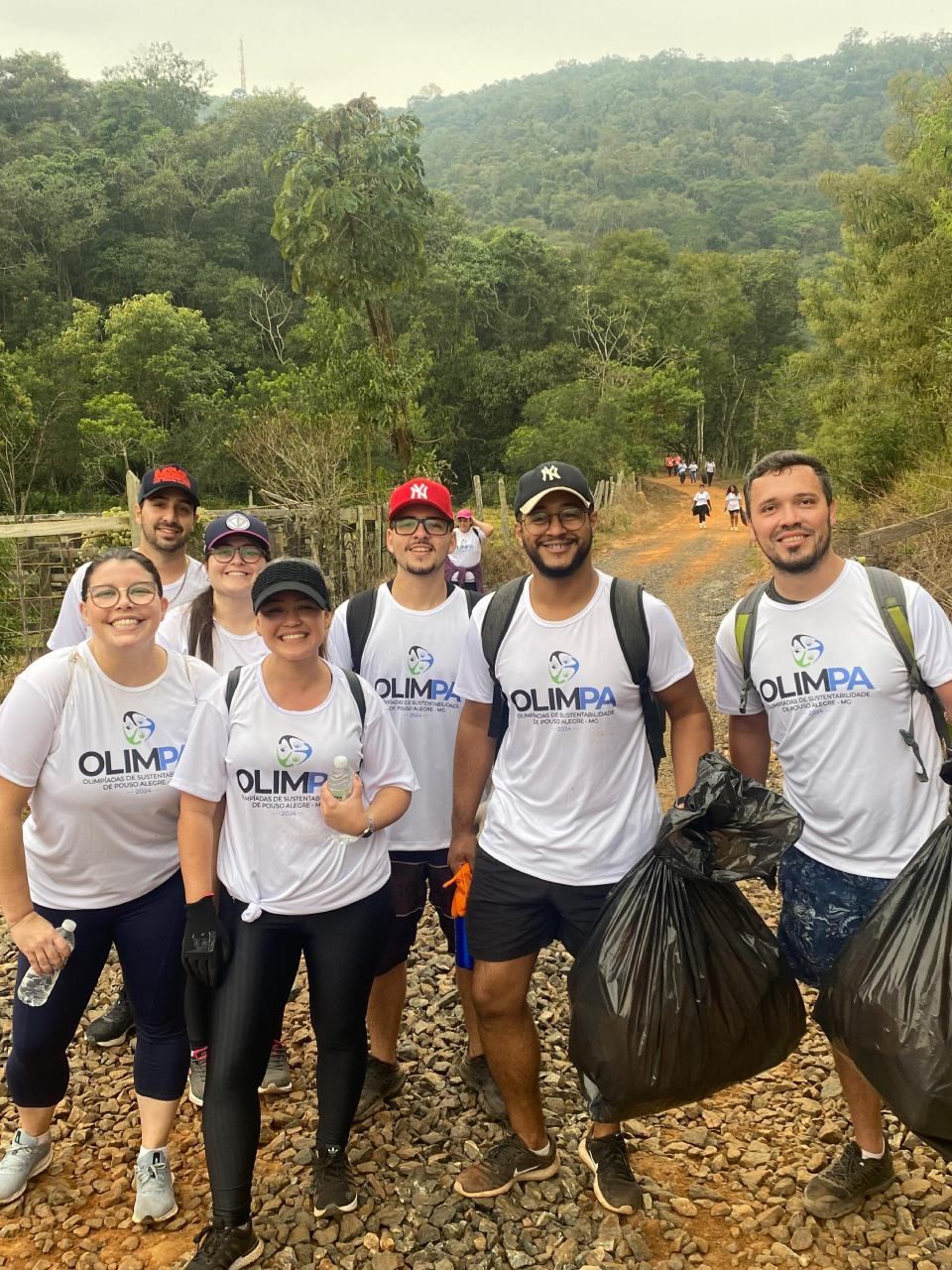 Um grupo de pessoas está de pé, uma ao lado da outra, em uma estrada de terra, segurando sacos de lixo.