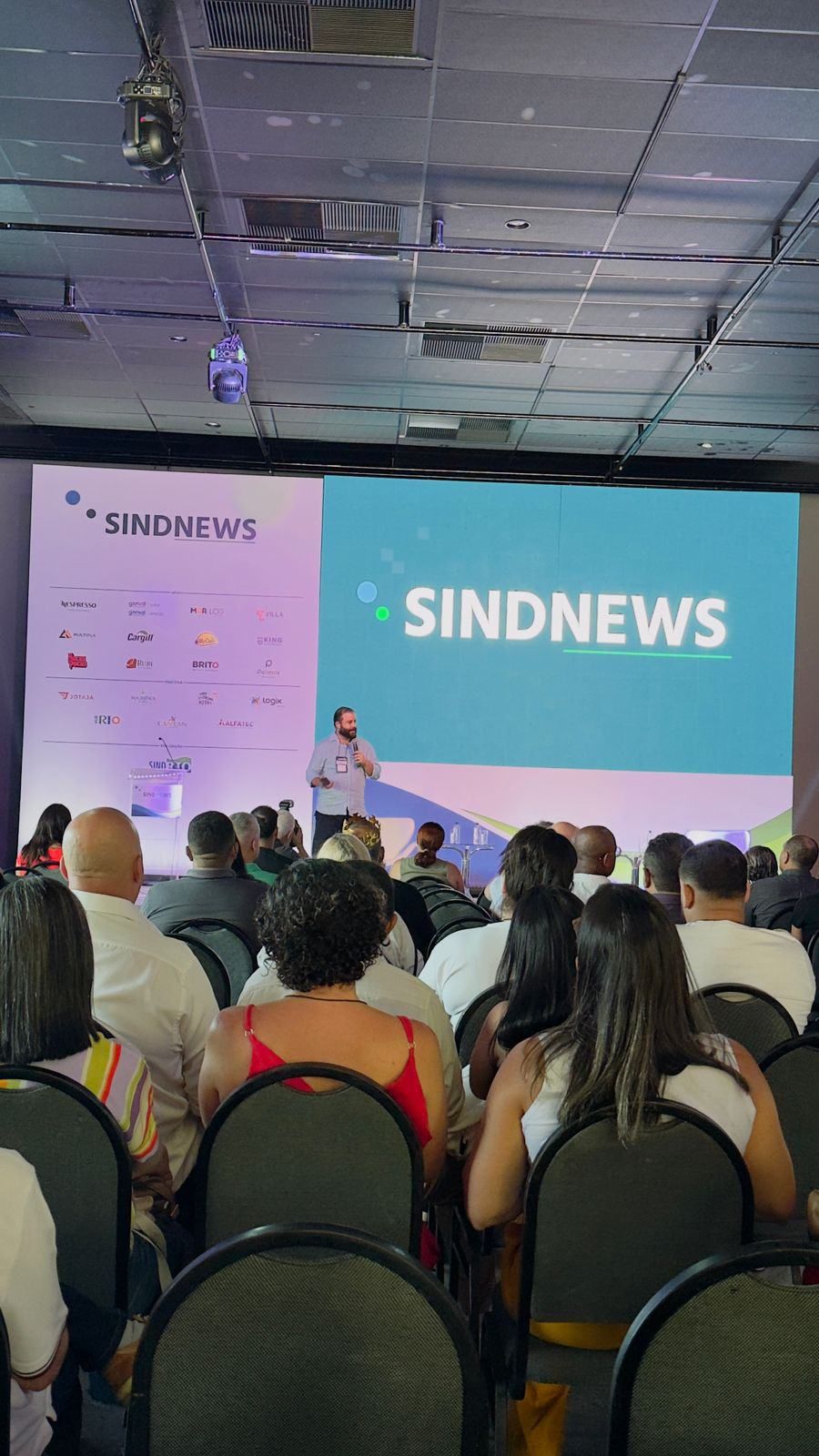 Um grupo de pessoas está sentado em cadeiras em frente a uma tela grande que diz sindnews.