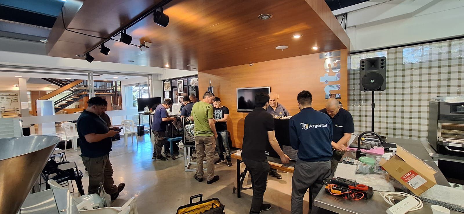 Pessoas se reuniram em um espaço interno com teto de madeira, algumas olhando para uma tela de TV e outras trabalhando em projetos.