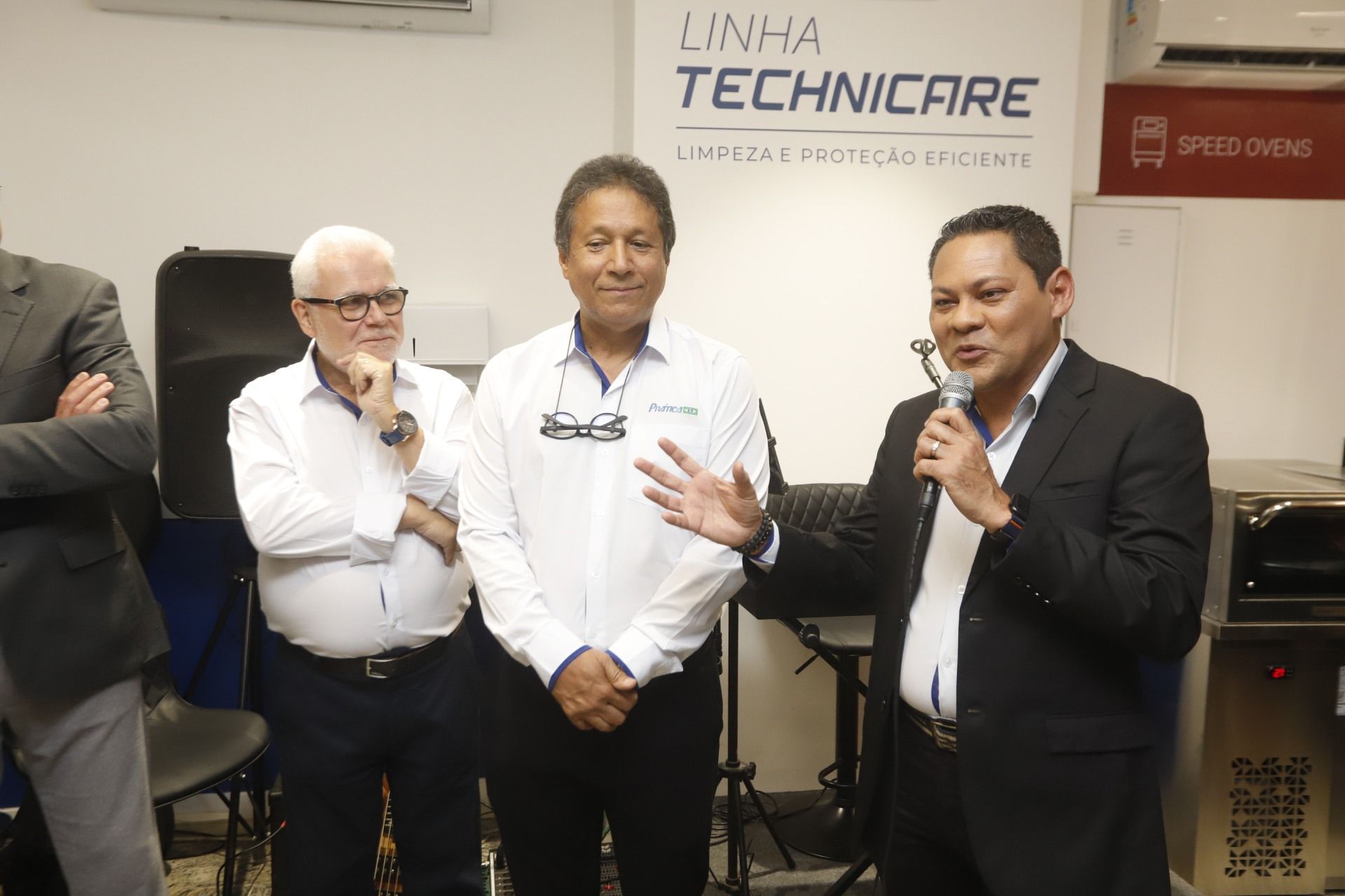 Três homens em um evento empresarial: um fala ao microfone, os outros observam. A placa diz 