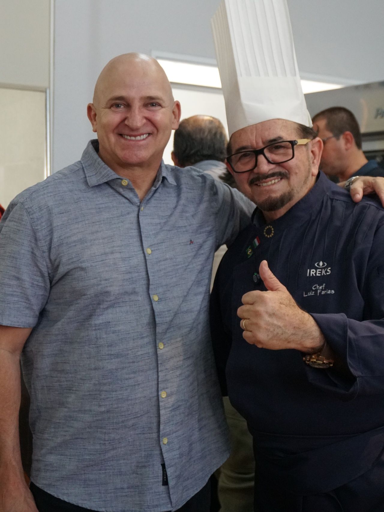 Dois homens sorrindo, um com chapéu de chef e uniforme fazendo sinal de positivo, o outro com camisa azul.