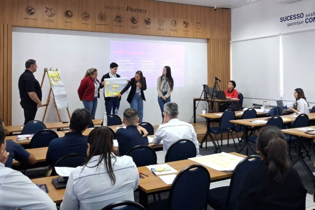Pessoas apresentando em uma conferência. Participantes sentados. Quadro de apresentação e tela visíveis. Cores neutras.