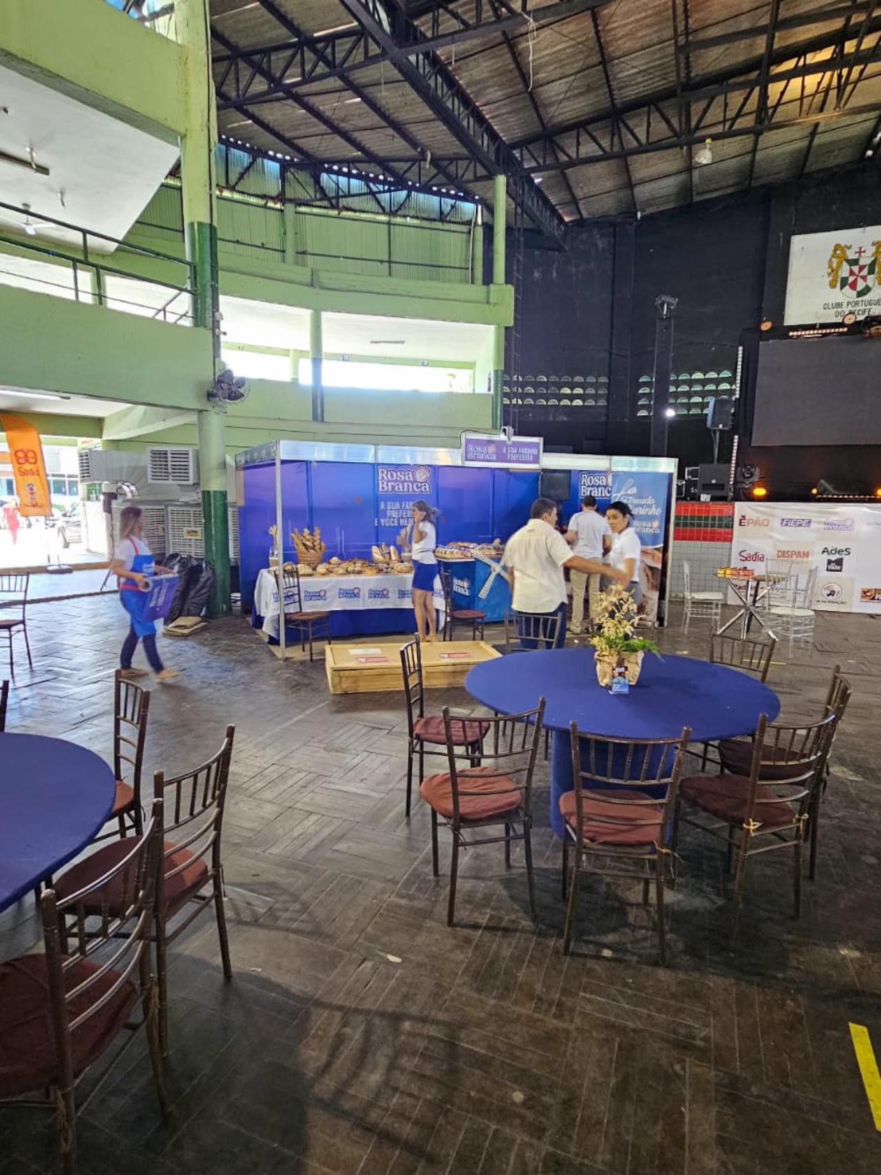 Um grupo de pessoas está de pé ao redor de mesas e cadeiras em uma sala grande.