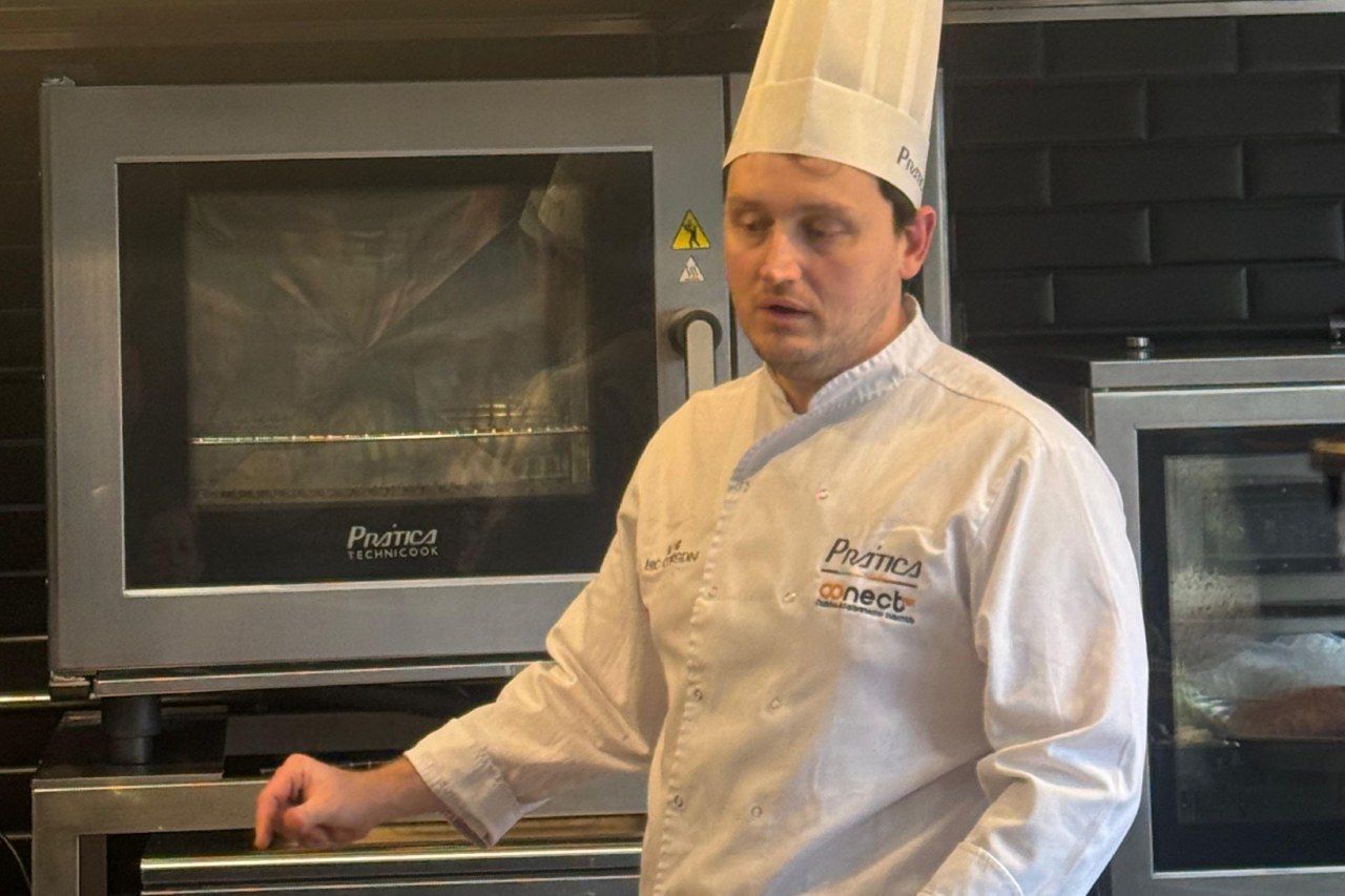 Chef de uniforme branco e chapéu, em pé em frente ao forno na cozinha, apontando com a mão.