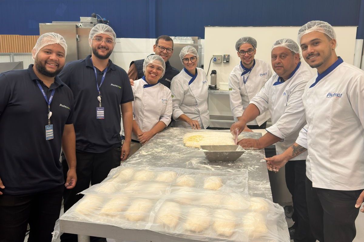 Grupo de pessoas em uma padaria, trabalhando na massa. Vestem uniformes de chef e toucas de cabelo, sorrindo.