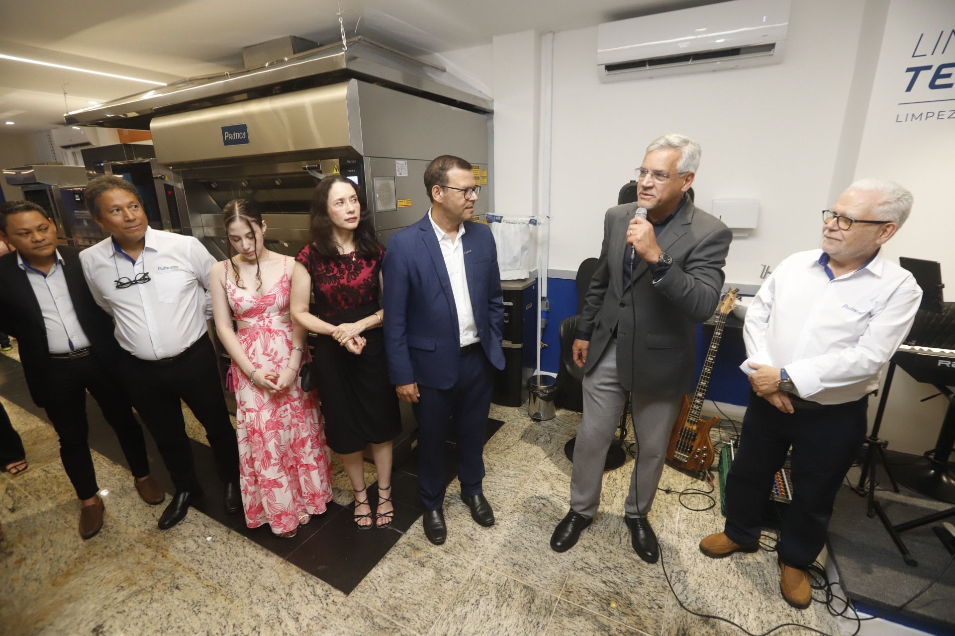 Pessoas em trajes formais, provavelmente em um evento. Um homem fala ao microfone. Equipamentos de cozinha ao fundo.