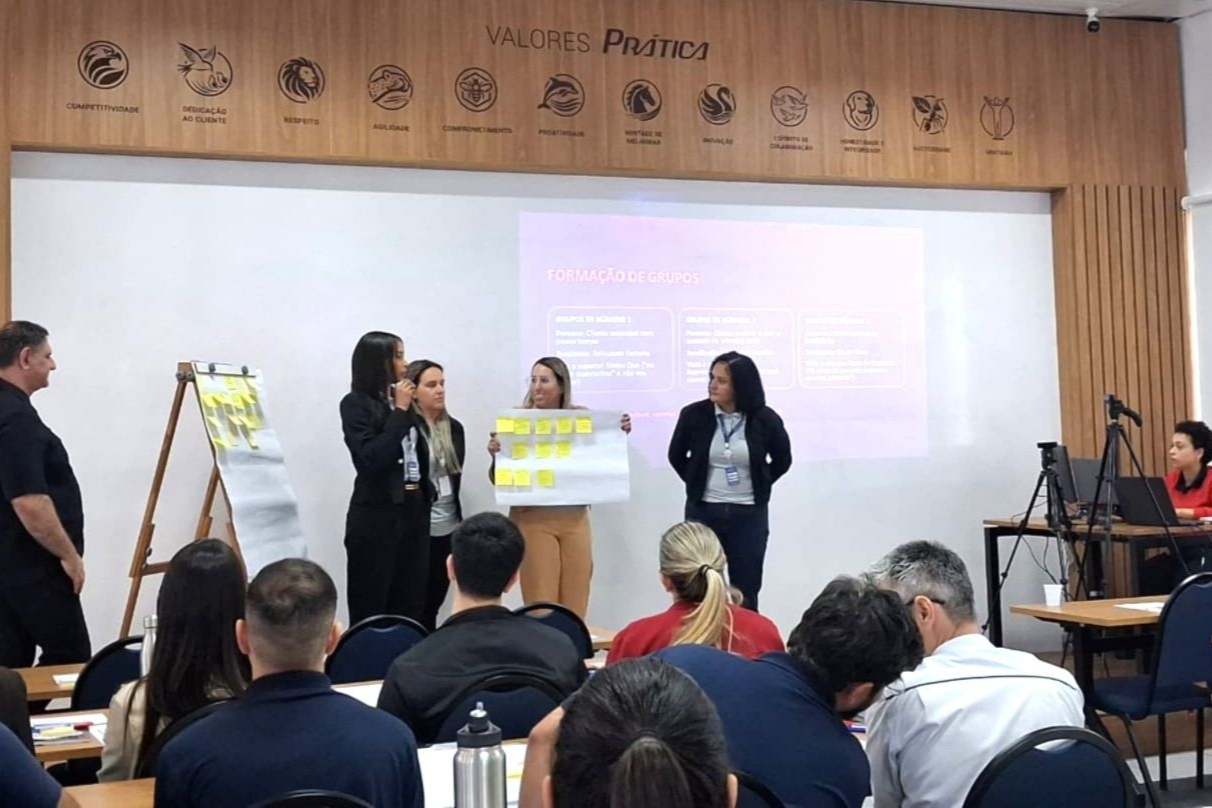 Uma apresentação em ambiente de workshop. Quatro mulheres apresentam um pôster. Público sentado.