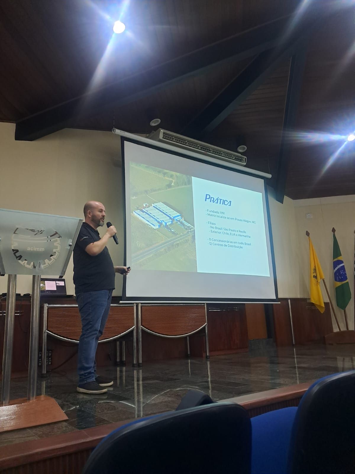 Um homem de camisa preta faz uma apresentação com slides. Ele está no palco de um auditório.