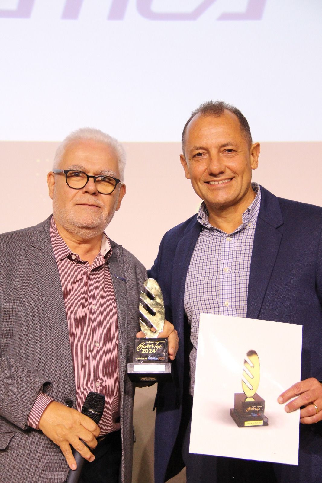 Dois homens estão de pé, um ao lado do outro, segurando troféus e um pedaço de papel.
