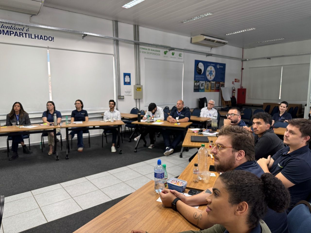 Um grupo de pessoas está sentado à mesa em uma sala de reuniões bem iluminada, assistindo a uma apresentação.