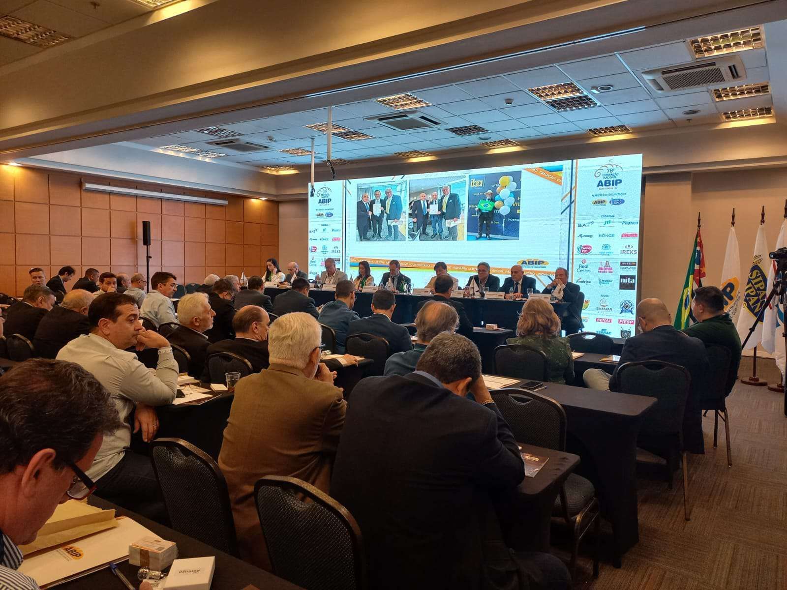 Um grande grupo de pessoas está sentado em mesas em uma sala de conferências.