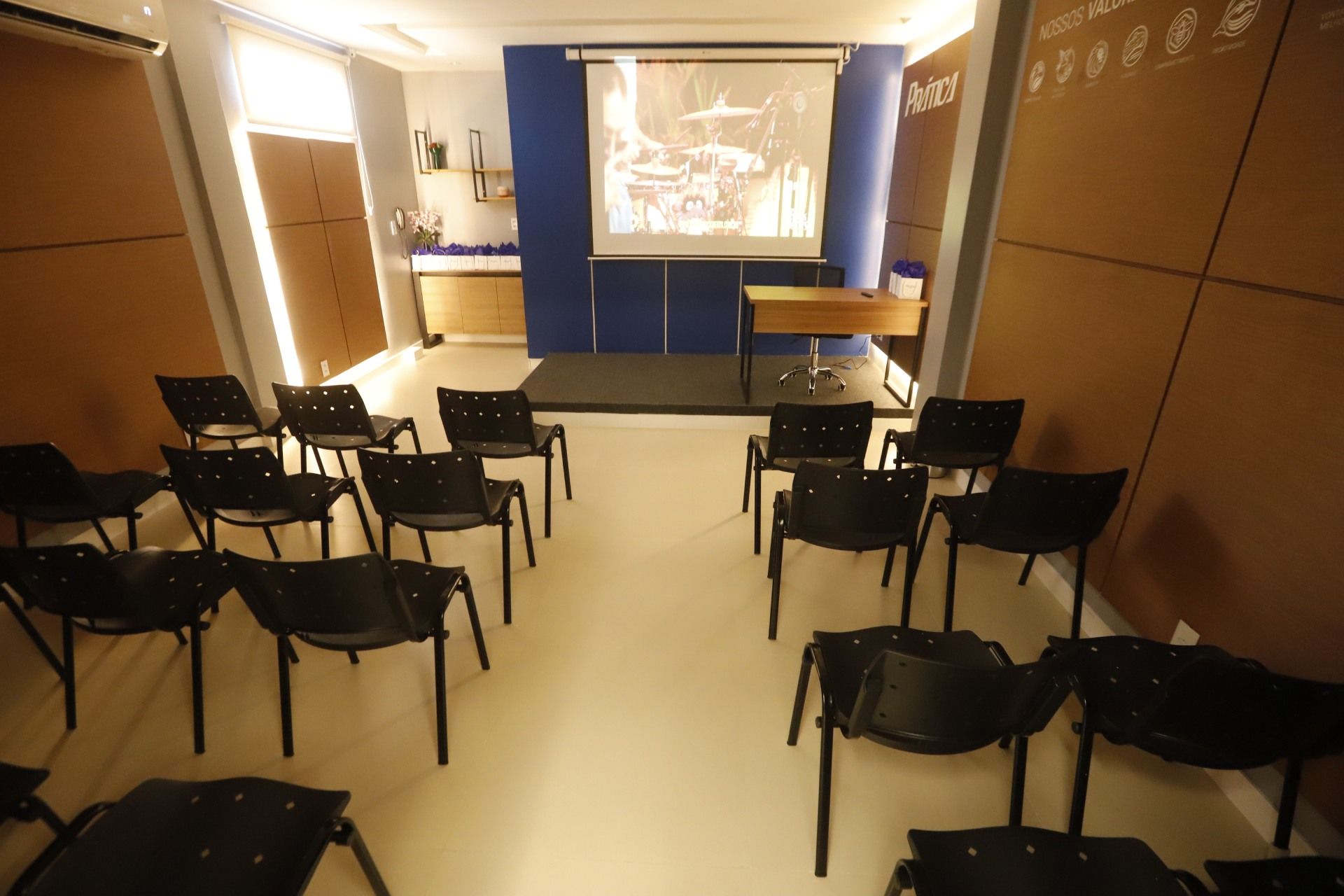 Sala de aula com fileiras de cadeiras pretas de frente para um palco com uma tela de projetor exibindo uma imagem em preto e branco.