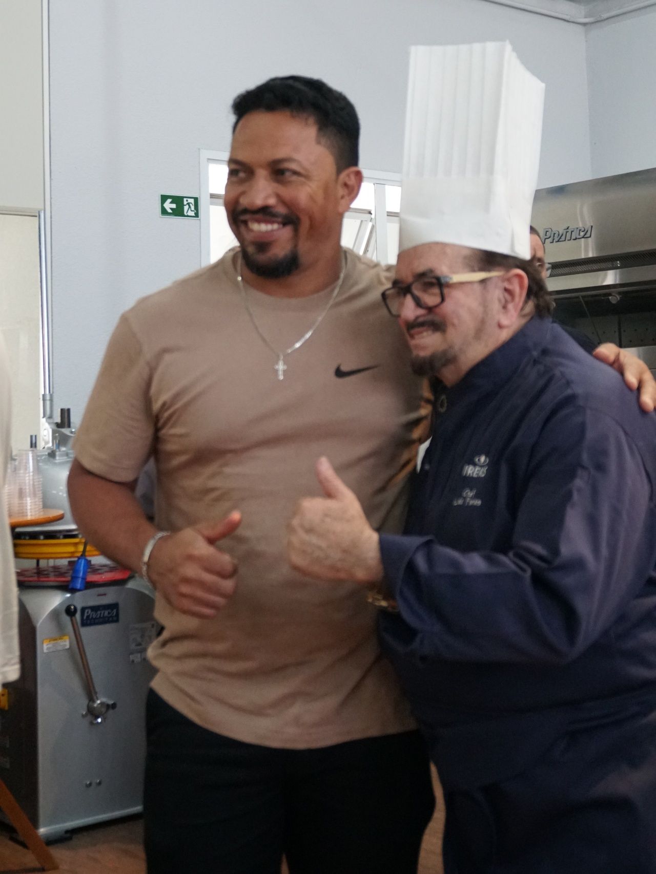 Homem de camisa bege e chef de terno azul posam, sorrindo e fazendo sinal de positivo em uma cozinha.