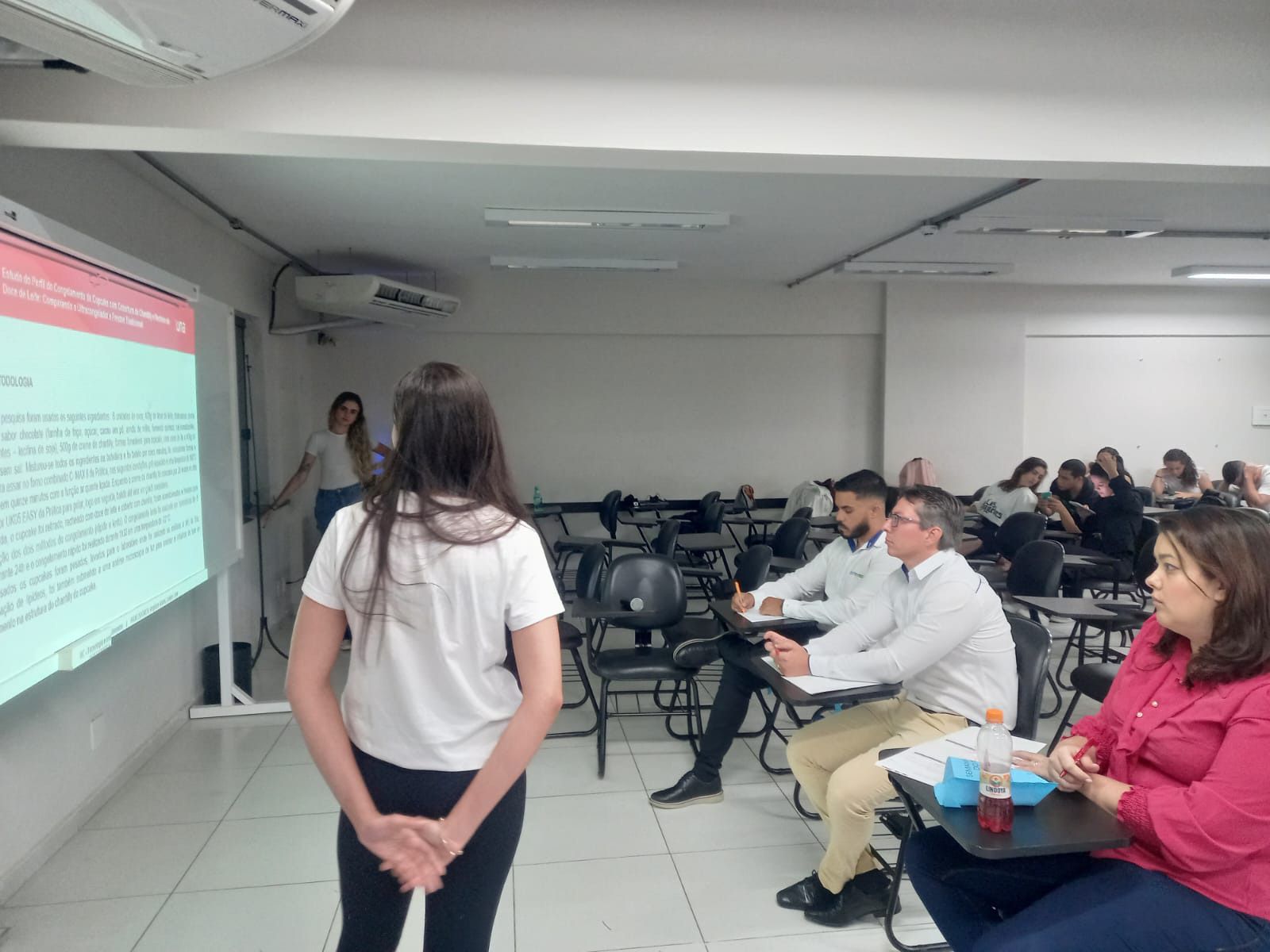 Uma mulher está parada na frente de um grupo de pessoas em uma sala de aula.