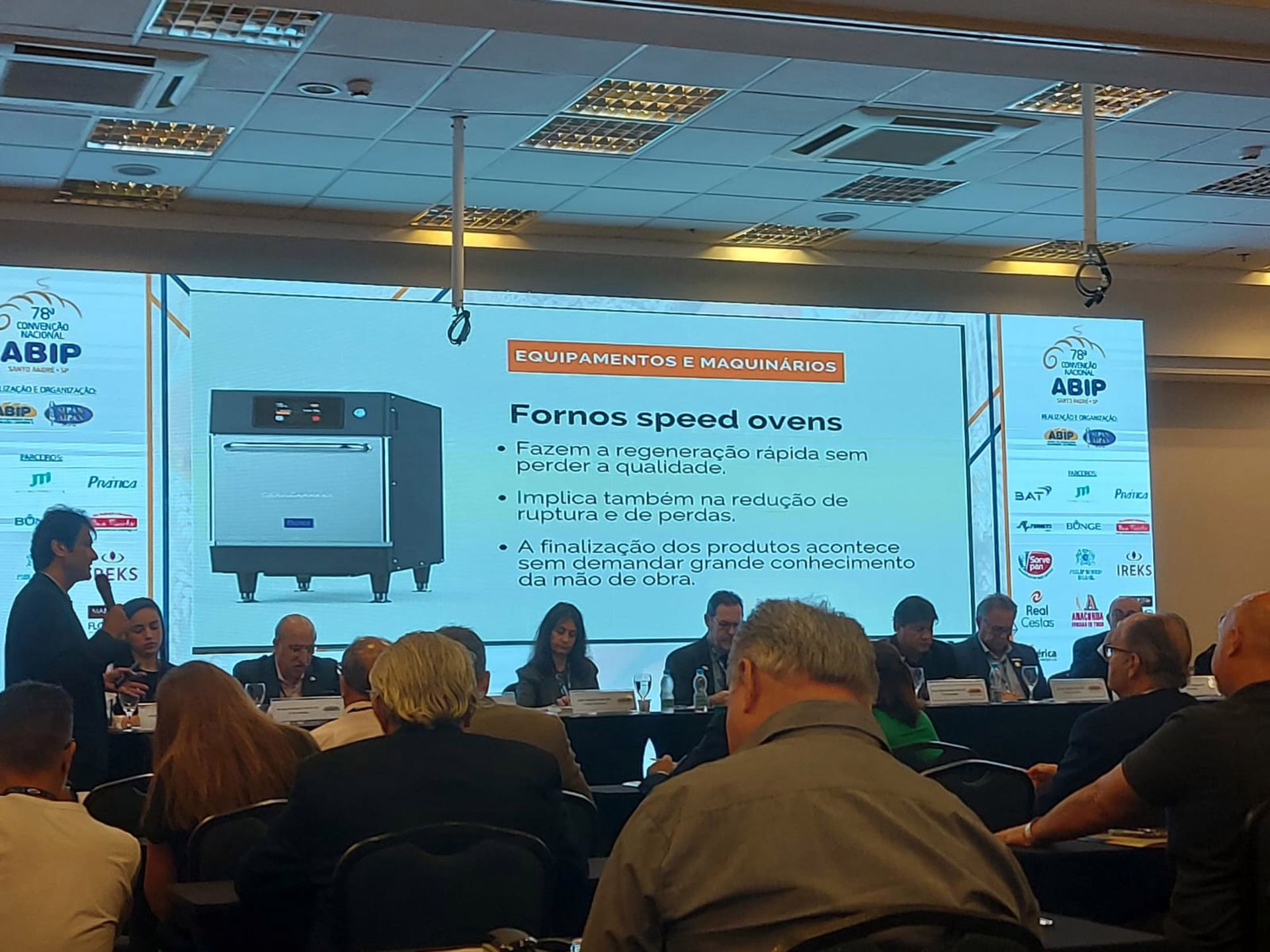 Um grupo de pessoas está sentado em frente a uma tela grande com a imagem de um forno